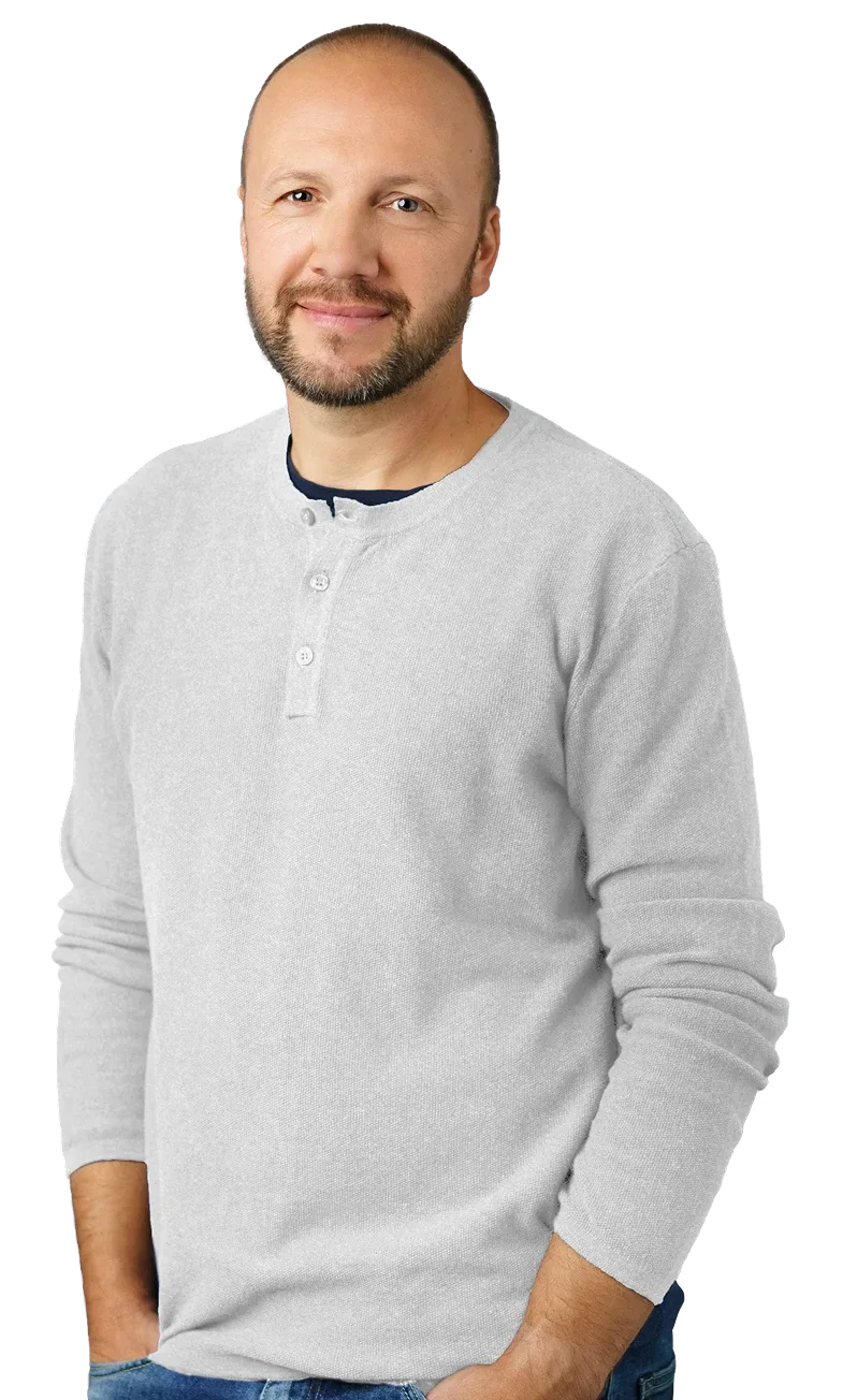 Man in a grey henley shirt and blue jeans standing with a slight lean, hands relaxed by his sides.